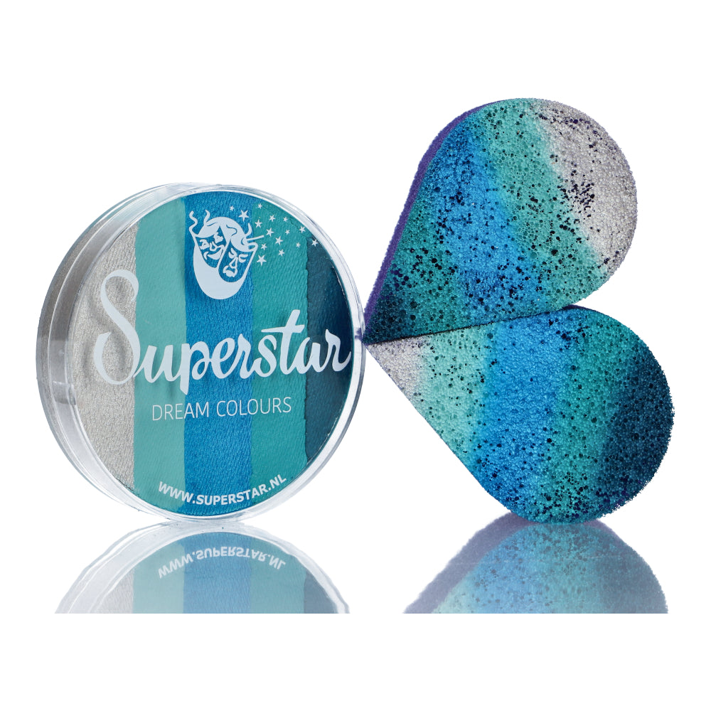 A round container of Split Cake Dream Colour Superstar - Ice (45gr) by Infinityfaces sits beside a heart-shaped sponge with a blue, teal, and gray gradient. The packaging has a star logo and complies with the European Cosmetics Directive. The sponge blends the vibrant hues beautifully.