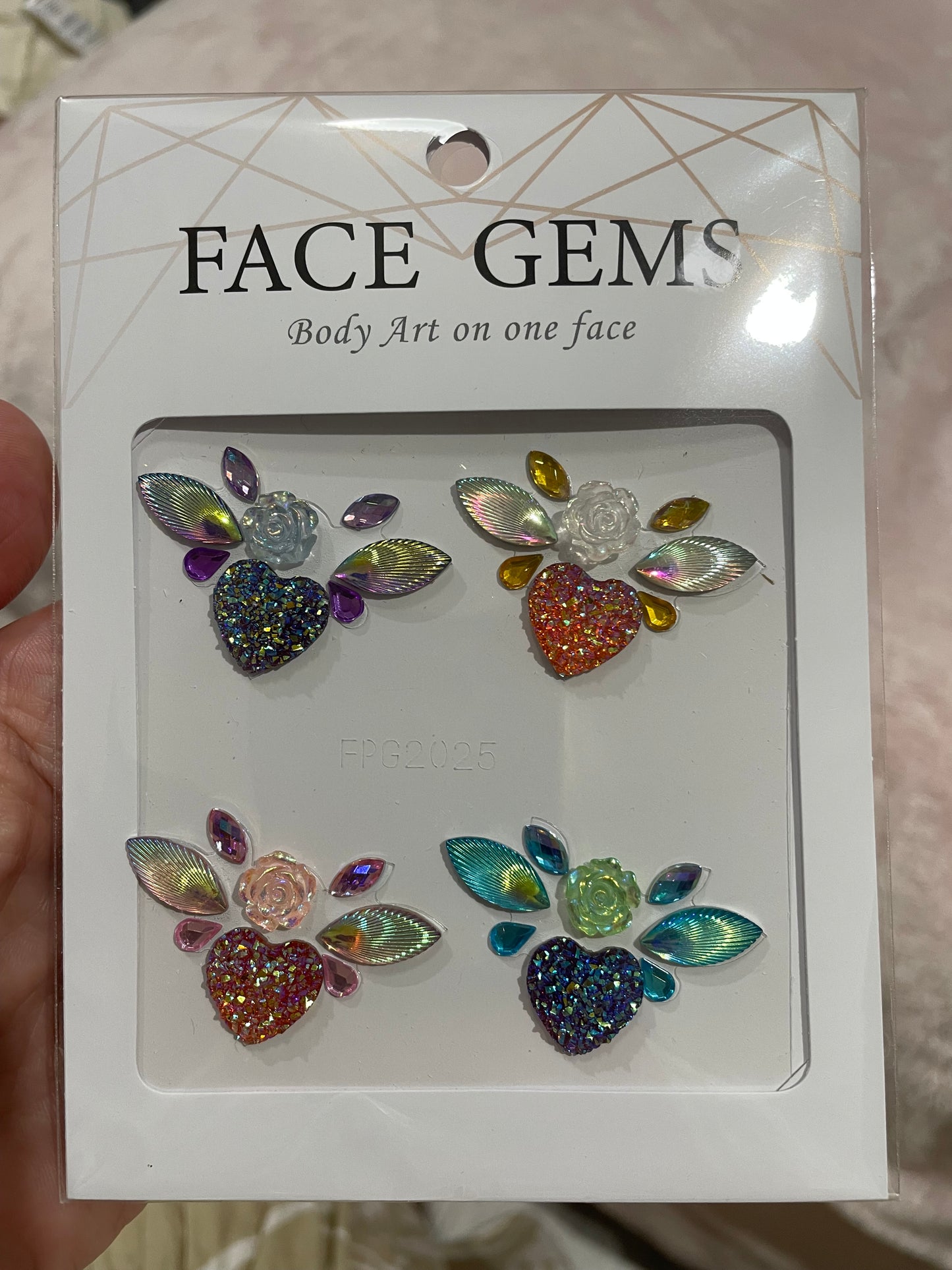 A hand holds a package of Infinityfaces Face- & Body Gems - Flower Heart. It features four heart-shaped gem designs with colorful leaves and flowers, ideal for skin embellishment. The label reads Face Gems: Body Art.