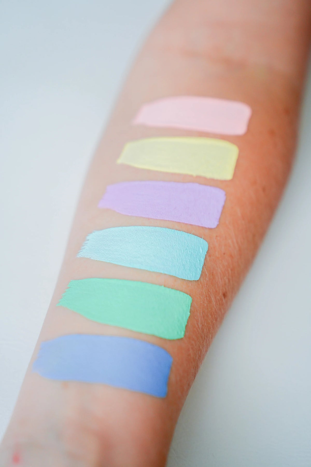 Six swatches of TAG Face & Body Paint - Pastel Turquoise (32g) along with pink, yellow, lavender, mint, and blue face paint applied in a vertical line on an arm.