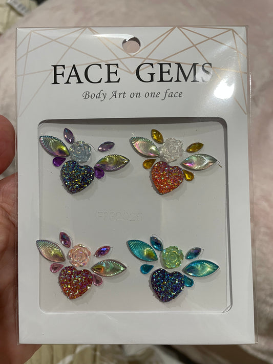 A hand holds a package of Infinityfaces Face- & Body Gems - Flower Heart. It features four heart-shaped gem designs with colorful leaves and flowers, ideal for skin embellishment. The label reads Face Gems: Body Art.