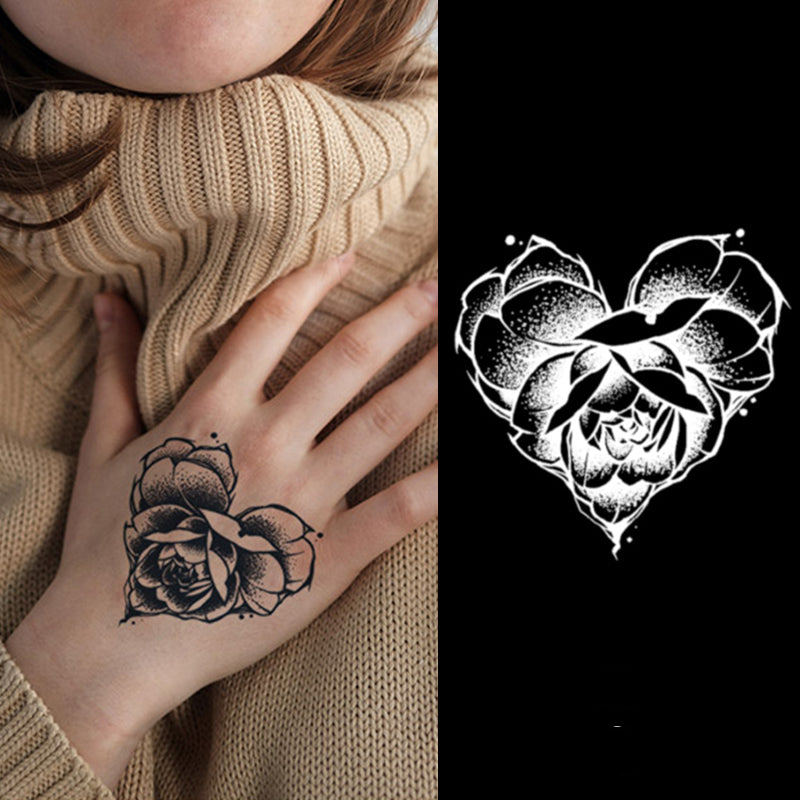 A person in a beige turtleneck displays the Temporary Herbal Tattoo - Fall in Love by Infinityfaces, featuring a black floral design on their hand. Beside it lies a white heart-shaped version on a black background.