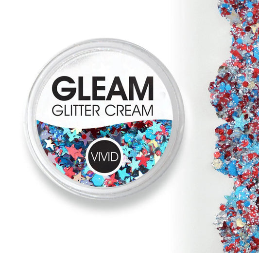 Round container labeled Infinity Faces Glitter Cream Gleam - Red White & Boom (10g) filled with colorful holographic glitter shapes like stars and hexagons in blue, red, and silver. Loose glitter is scattered on the right side.