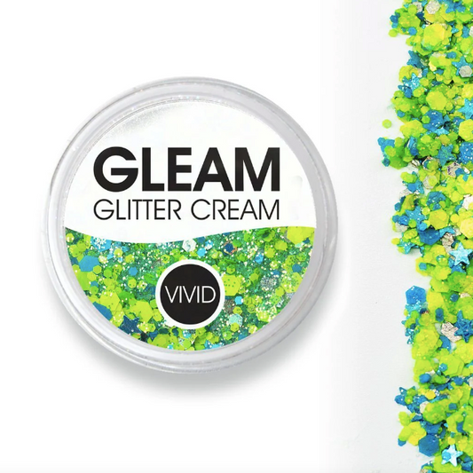 A round container labeled Infinity Faces Glitter Cream Gleam - Breeze (10g) holds blue and green glitter cream. Loose blue, green, and yellow glitter is striped on the white surface beside it.