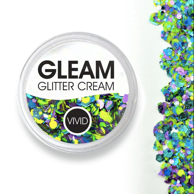 A round container of Infinity Faces Glitter Cream Gleam - Wild Bloom (10g) sits beside a scattered mix of chunky, colorful green, blue, and purple glitter on a white surface.