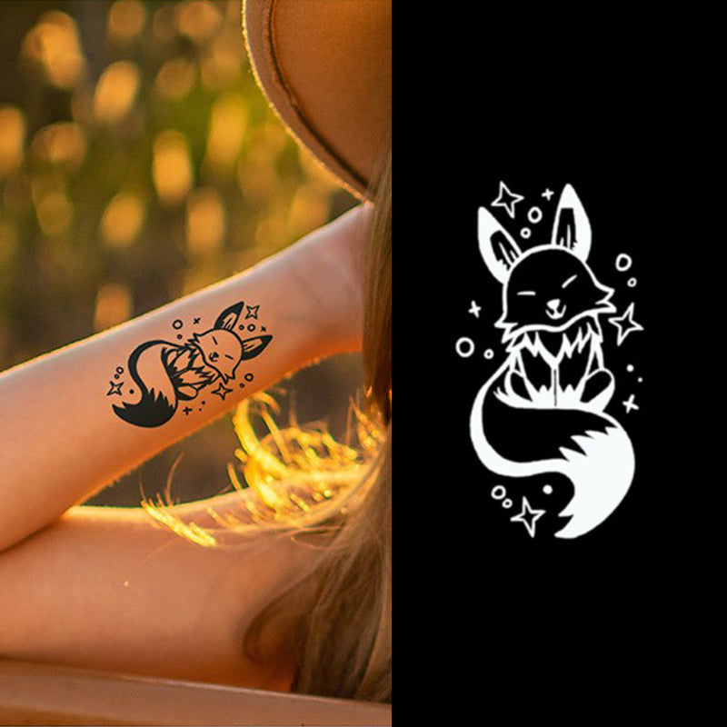 An individual with long hair sports a large hat, revealing their forearm adorned with the Infinityfaces Temporary Herbal Tattoo - Little Fox. It features a stylized mythical fox, artfully repeated in white on black, made with skin-safe plant-based ink lasting up to 14 days.