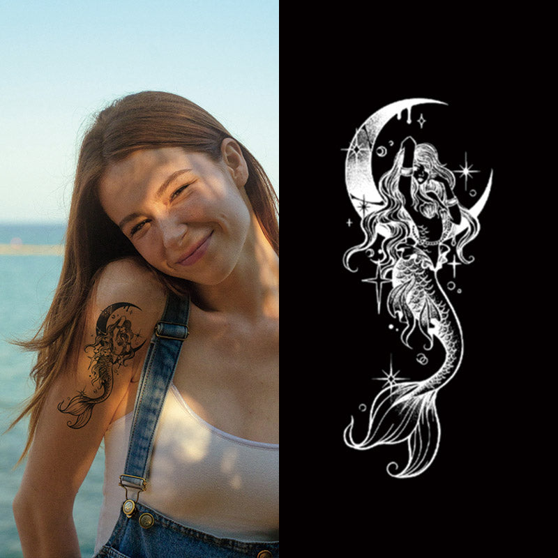 A woman with a smile displays her Temporary Herbal Tattoo - Mermaid from Infinityfaces, showcasing a floral design on her arm by the sea. Close by, an illustration of a long-haired mermaid sits on a crescent moon against a black backdrop.